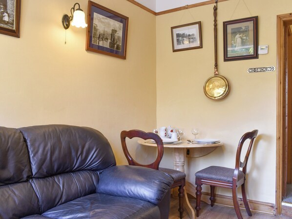 Une pièce avec un canapé en cuir, une table à manger dressée avec des assiettes, des verres et un pichet, et un panneau « DÉFENSE DE FUMER » accroché au mur.