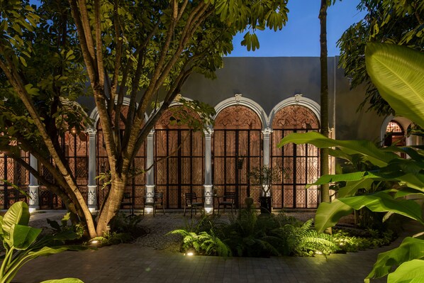Une cour intérieure avec un bâtiment doté d’arcades et de colonnes, entourée d’une végétation luxuriante et d’arbres.
