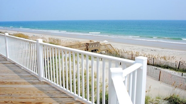 Eine Holzdielen-Terrasse mit weißen Geländern, die auf einen Strand mit einer Holzpromenade und einem Holzgebäude blickt.