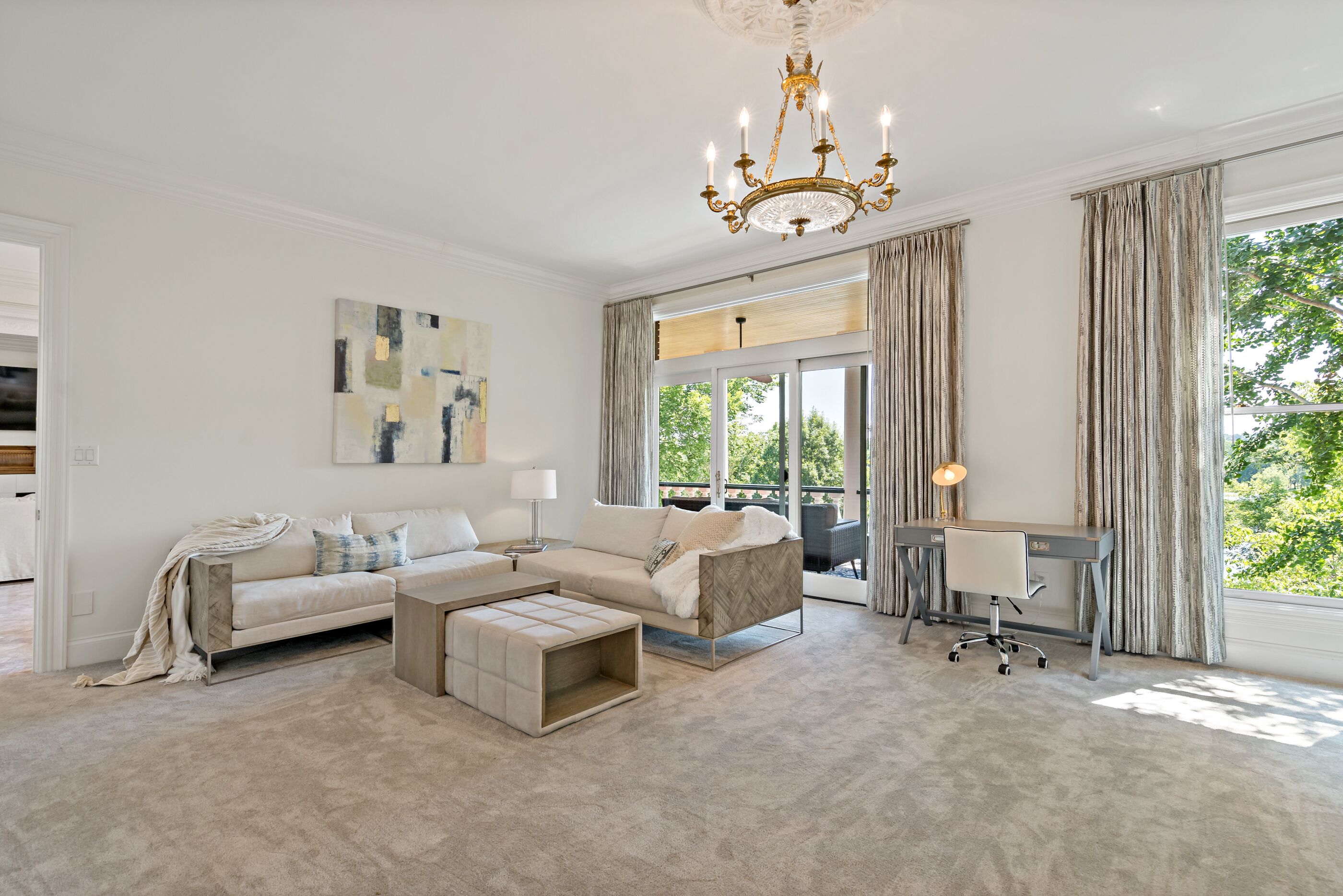 Un salon spacieux doté d’une grande fenêtre, d’un lustre, d’un ensemble de canapés, d’une table basse et d’un bureau avec une chaise.