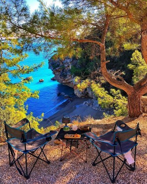 A table with camping gear and two chairs overlooking a beach with clear blue water and rocky cliffs.