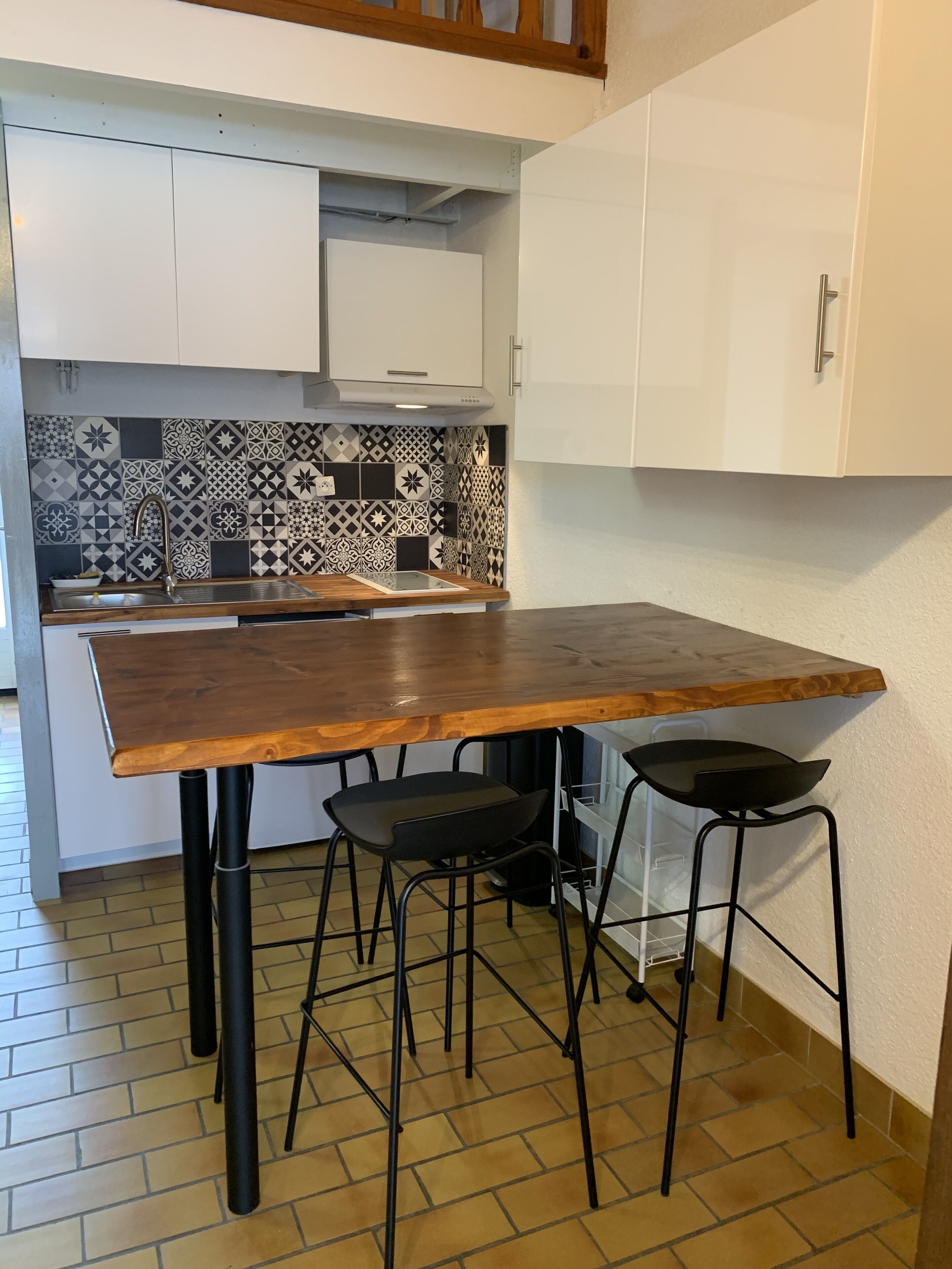 Een keuken met een houten tafel, twee zwarte krukken, een betegelde achterwand en witte kasten.
