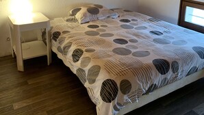 Ein Schlafzimmer mit Holzboden, einem weißen Bett mit gemustertem Überwurf, einem Nachttisch mit Lampe und einem Fenster.