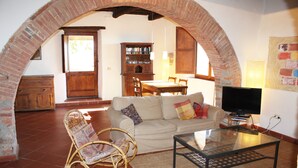 A living room with a brick archway, a sofa, a glass coffee table, a wicker chair, a wooden cabinet, and a television. - Traumblick in die Toskana - Appartment mit Pool auf Hügel zw. Siena u. Florenz (Toscana)