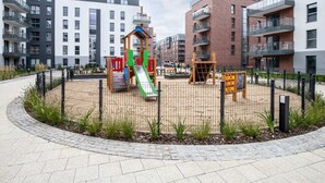 A playground enclosed by a fence with a slide and climbing structures, surrounded by a paved walkway and greenery.