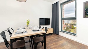 A modern dining area with a wooden table set for four, black chairs, a flat-screen TV, and a large window with a view of the city.