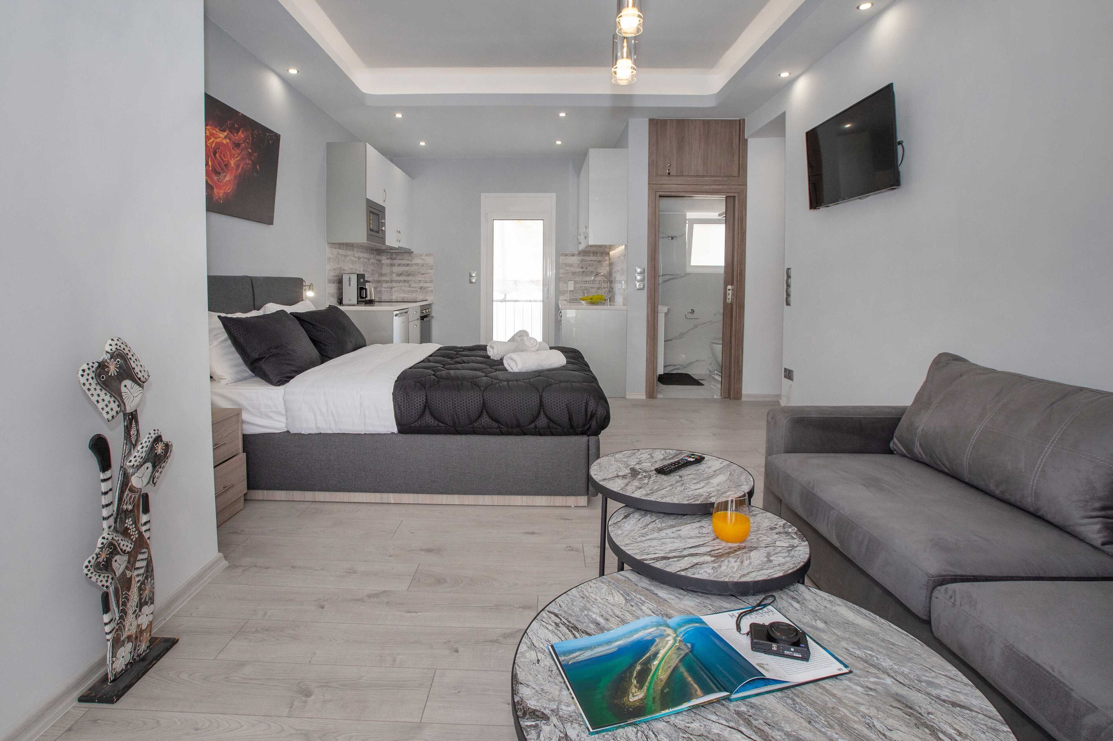 A modern hotel room with a bed, a sofa, a marble coffee table, and a small kitchen area.