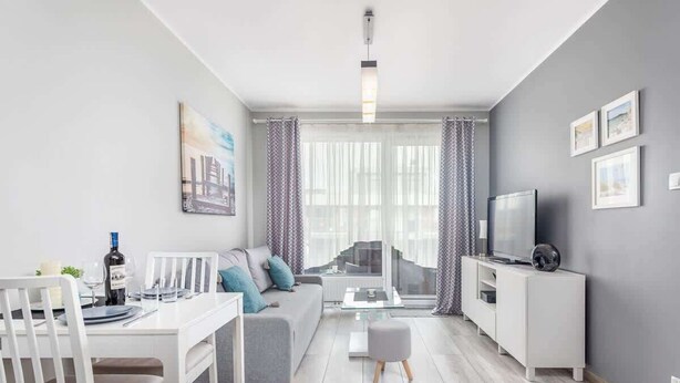 A modern living room with a dining area, grey sofa, white TV stand, and a large window with curtains.