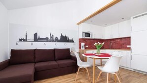 Una sala de estar moderna con un sofá marrón, una mesa de comedor con sillas blancas y una zona de cocina con armarios blancos y un revoque de ladrillo rojo.