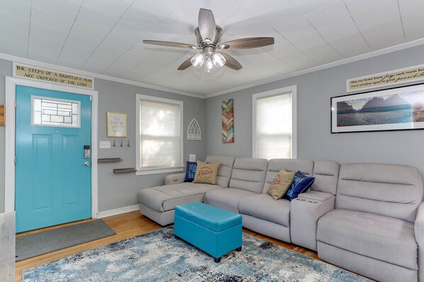 A living room with a teal door, a ceiling fan, a grey sofa with cushions, a blue ottoman, and a patterned rug.