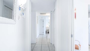 Un pasillo luminoso y blanco con suelo de baldosas que conduce a una habitación con cama y sillón.
