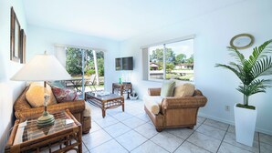 Un salón luminoso con muebles de mimbre, un televisor de pantalla plana y ventanas amplias que ofrecen vistas al exterior.