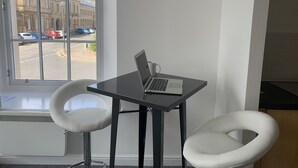 Une pièce avec une fenêtre donnant sur la rue, une table avec un ordinateur portable et deux chaises blanches.