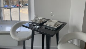 Une table à manger avec deux assiettes blanches, un verre à vin et une fourchette. La table est noire et se trouve à côté d’une fenêtre donnant sur une rue et des immeubles.