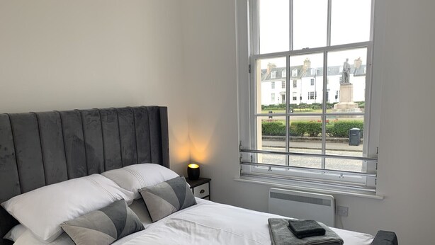 Une chambre à coucher avec un grand lit, une table de chevet avec une lampe et une fenêtre donnant sur des immeubles.