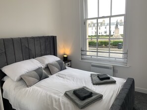A bedroom with a large bed, a nightstand with a lamp, and a window with a view of buildings. - Ayr Cozy Corner Apartment (Ayr)