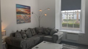 Un salon avec un canapé gris, une table basse blanche, un tableau représentant un chien et une fenêtre donnant sur des immeubles.