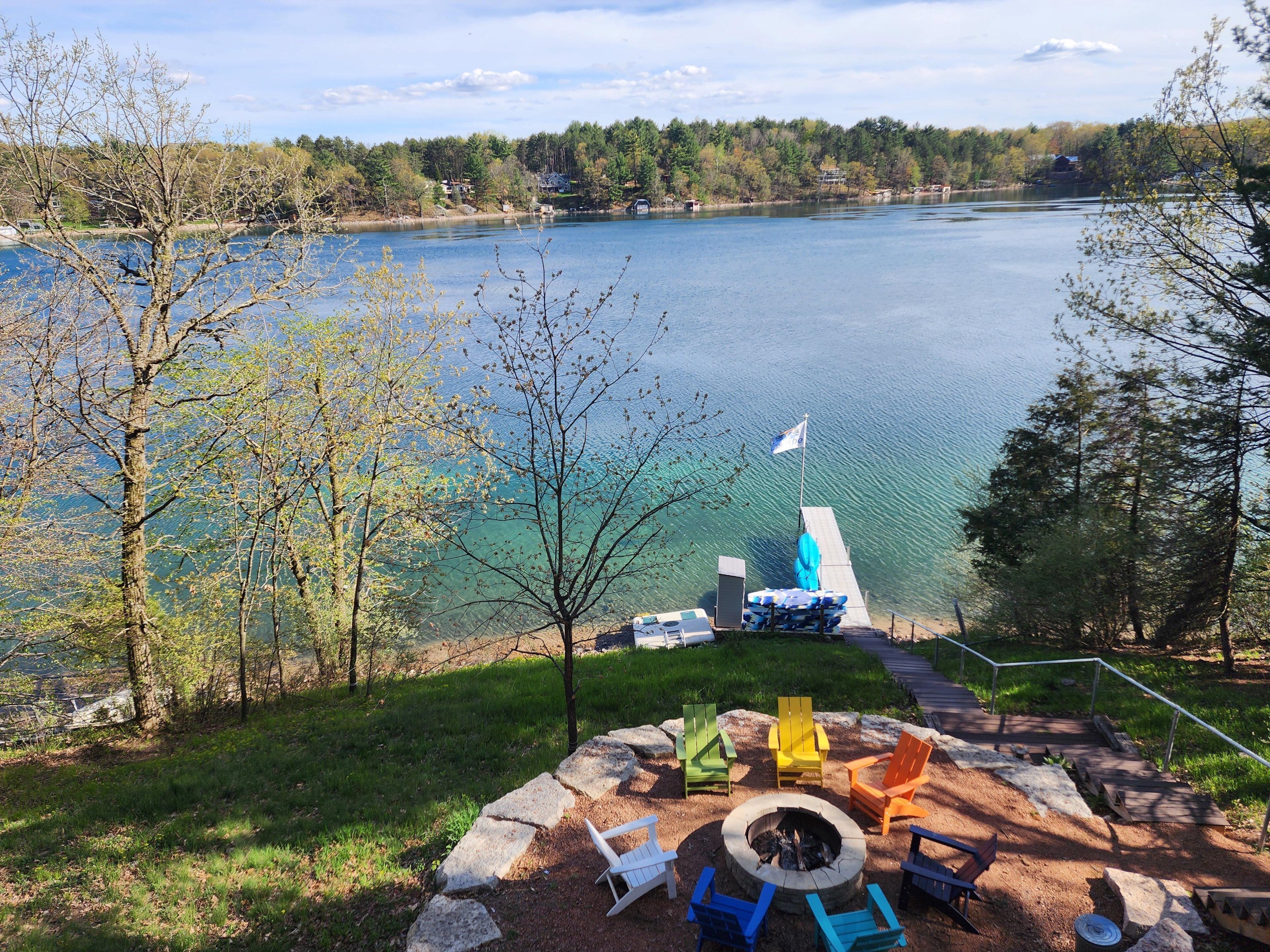 A lakeside view with a dock, a flag, and a fire pit area with Adirondack chairs.