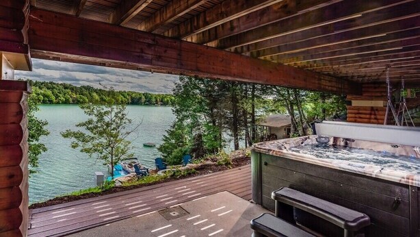 Une terrasse en bois avec un jacuzzi, donnant sur un lac où l’on peut voir des bateaux et une végétation luxuriante.