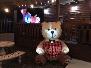 A large inflatable bear with a heart-shaped balloon in front of a wooden cabin with a television displaying colorful lights. - Poblado Log Cabin Camper  (Boquerón)