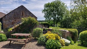A stone building with a sloped roof, surrounded by a well-maintained garden with various plants and a picnic table. - Gorgeous Stone Cottage with tasteful interior (Chathill)