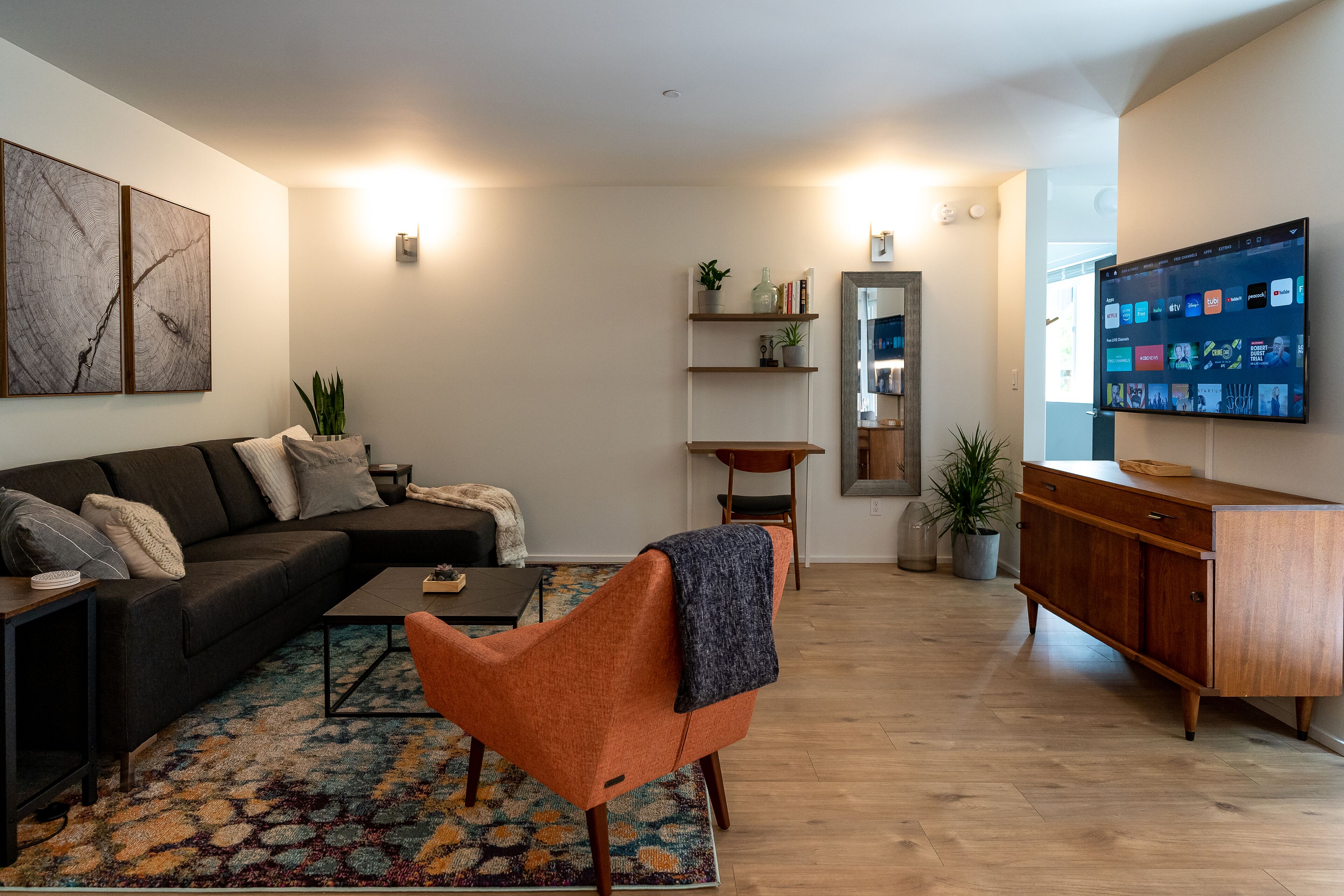 Un salon moderne comprenant un canapé d’angle, une table basse, un fauteuil orange, un téléviseur fixé au mur et un meuble en bois.