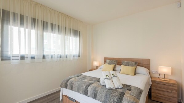 Una habitación de hotel con cama, dos mesitas de noche, ventana con cortinas y mesita de noche con lámpara.