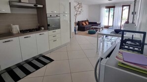 A modern kitchen with white cabinets, a gas stove, and a black and white striped mat. There is a dining area with a table and chairs, and a sofa in the background. - Villa Luigi in Poggio dei Pini: villa accommodation with parking and wallbox (Capoterra)