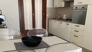A modern kitchen with white cabinets, a stove, and a dining area with a white table and clear chairs. - Villa Luigi in Poggio dei Pini: villa accommodation with parking and wallbox (Capoterra)