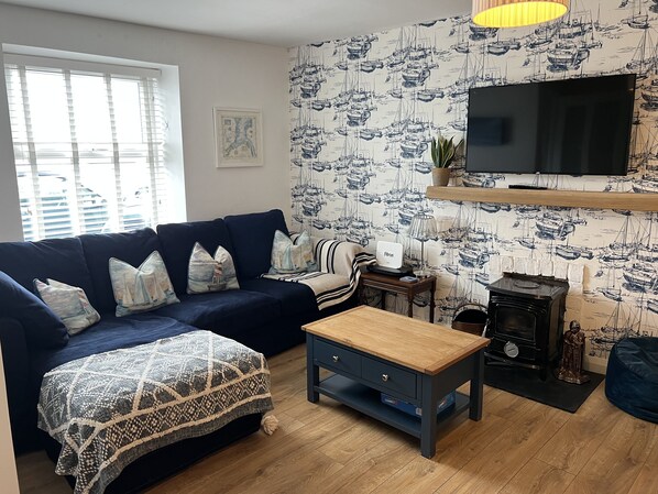 A living room with a dark blue sofa, patterned cushions, a wooden coffee table, a television mounted on the wall, and a patterned wallpaper.