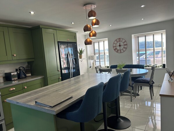 A modern kitchen with green cabinets, a stainless steel refrigerator, and a wooden island with bar stools. There is a dining area with a round table and chairs, and a large window with blinds.
