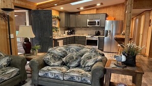 A cozy living room with a green sofa and matching armchair, a wooden coffee table with a duck decoration, and a kitchen with modern appliances and wooden cabinetry. - Whispering Pines Lakeside Retreat  (Park Rapids)