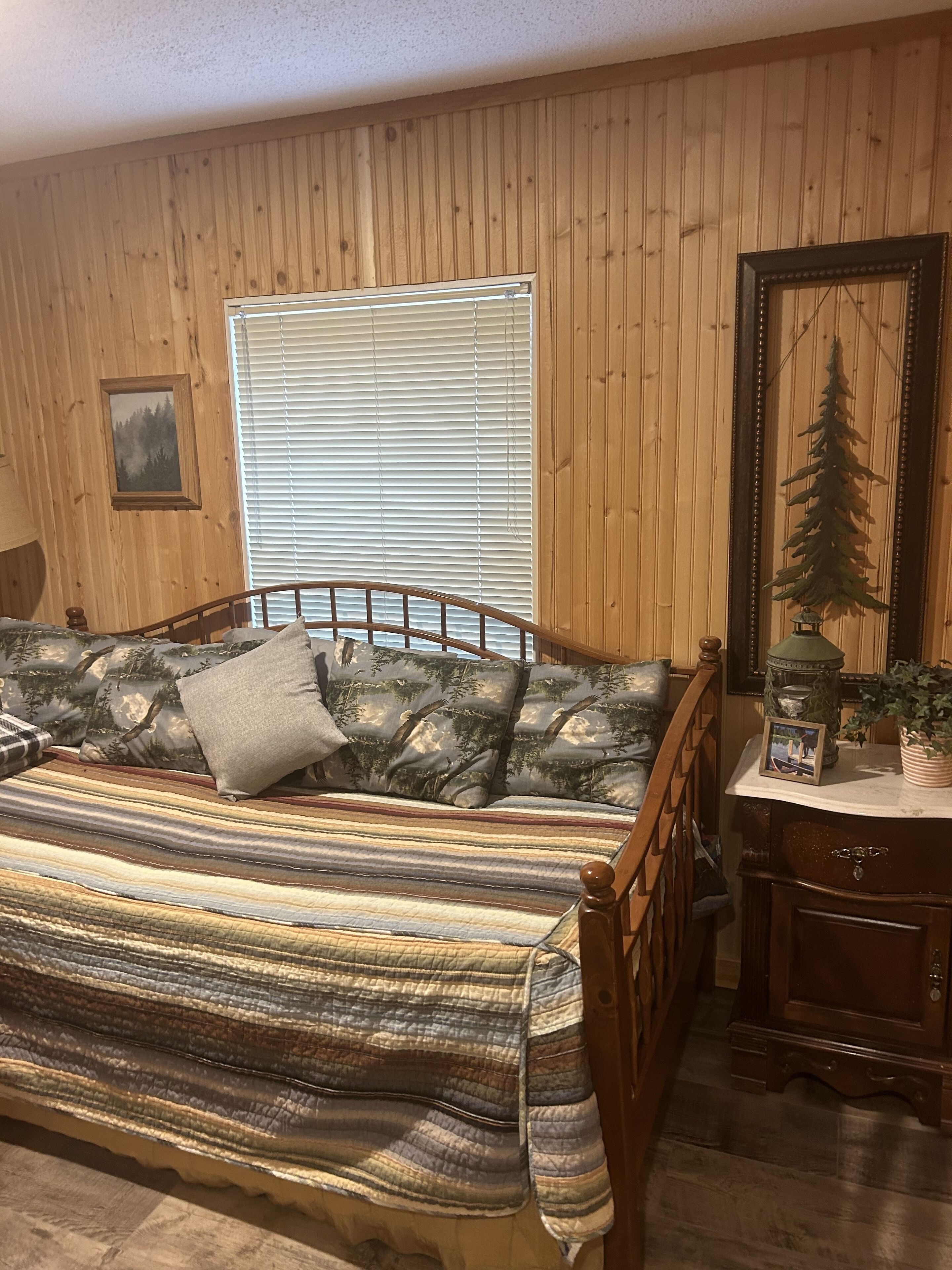 Une pièce chaleureuse avec un mur lambrissé en bois, un lit recouvert d’une couverture à rayures, une fenêtre avec des stores, un tableau encadré et une petite table sur laquelle est posée une plante.