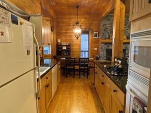 A wooden interior with a kitchen area featuring a dining table, chairs, a refrigerator, and a stove. - Breathtaking Mountain Views • Wraparound Deck • Fire Pit • Game Room (Tuckasegee)