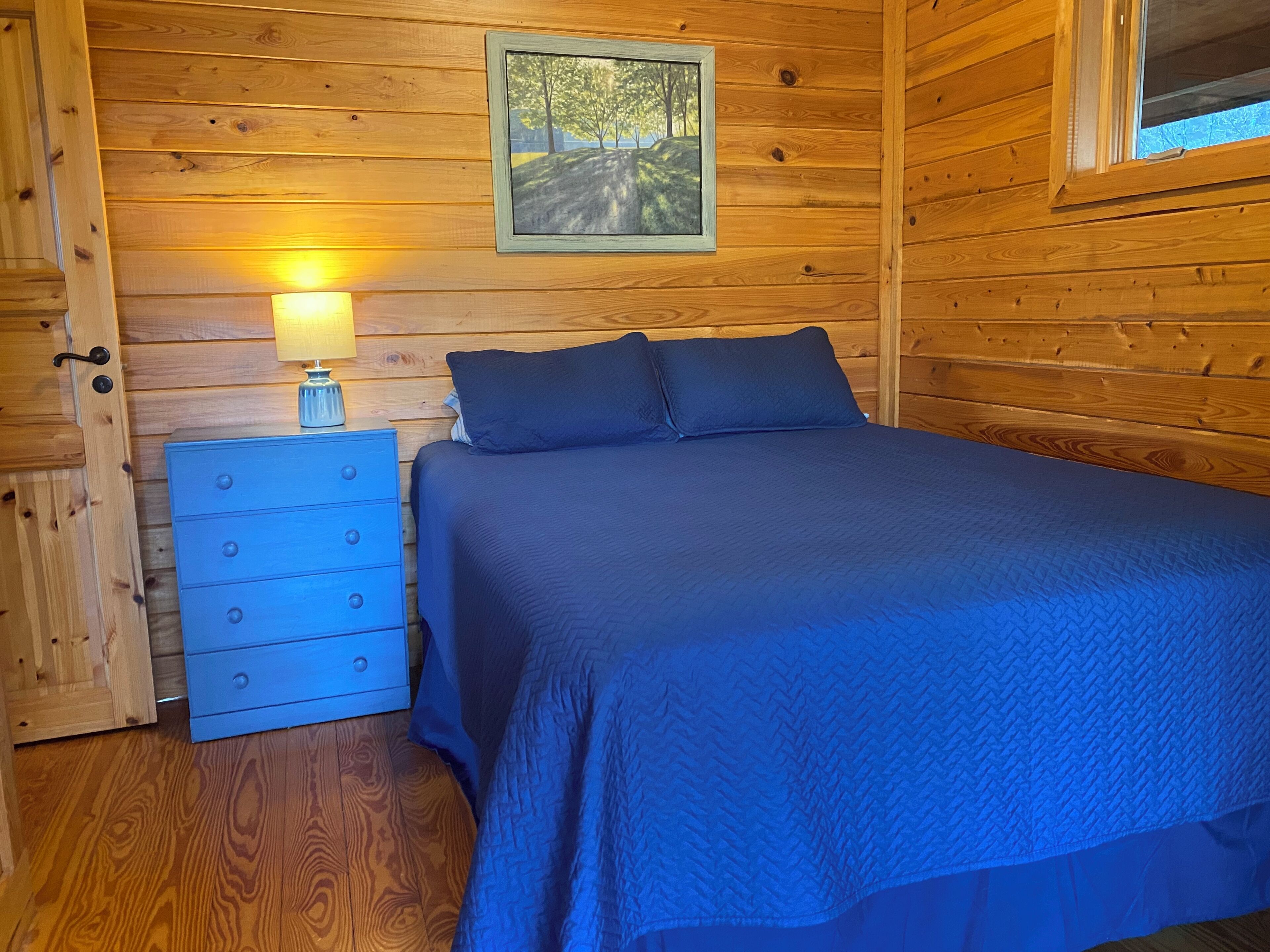 A wooden cabin bedroom with a bed, dresser, and a painting on the wall.