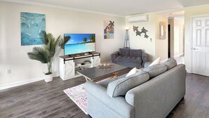 A living room with a grey sofa, a coffee table, a flat-screen TV, a potted plant, and a painting of a beach scene.