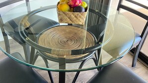 A round glass table with a woven placemat, surrounded by black chairs with woven bases. A woven basket with fruits is placed on the table.