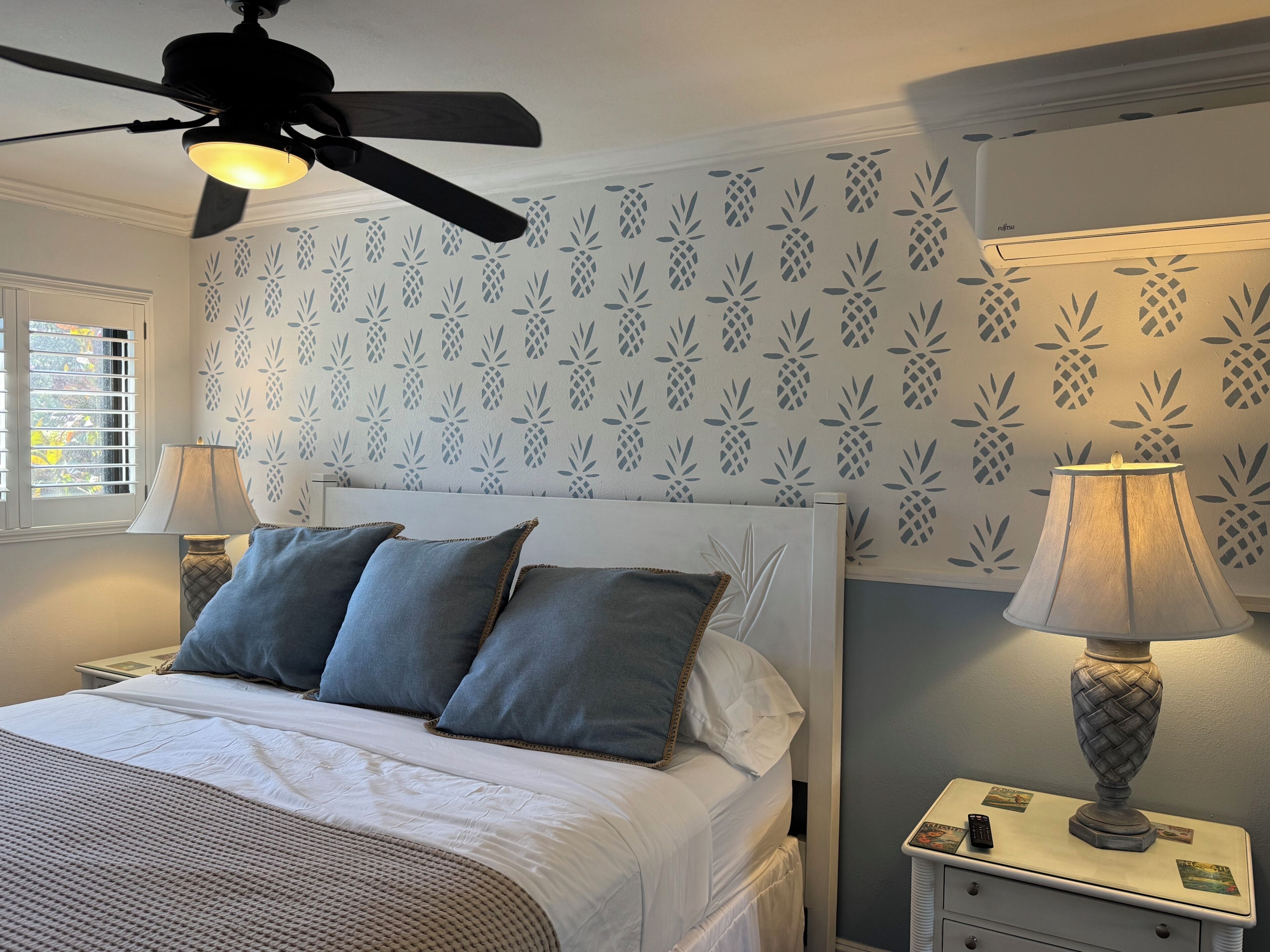 Une chambre à coucher avec un lit, un ventilateur de plafond, une table de chevet avec une lampe et un mur orné d’un motif ananas.