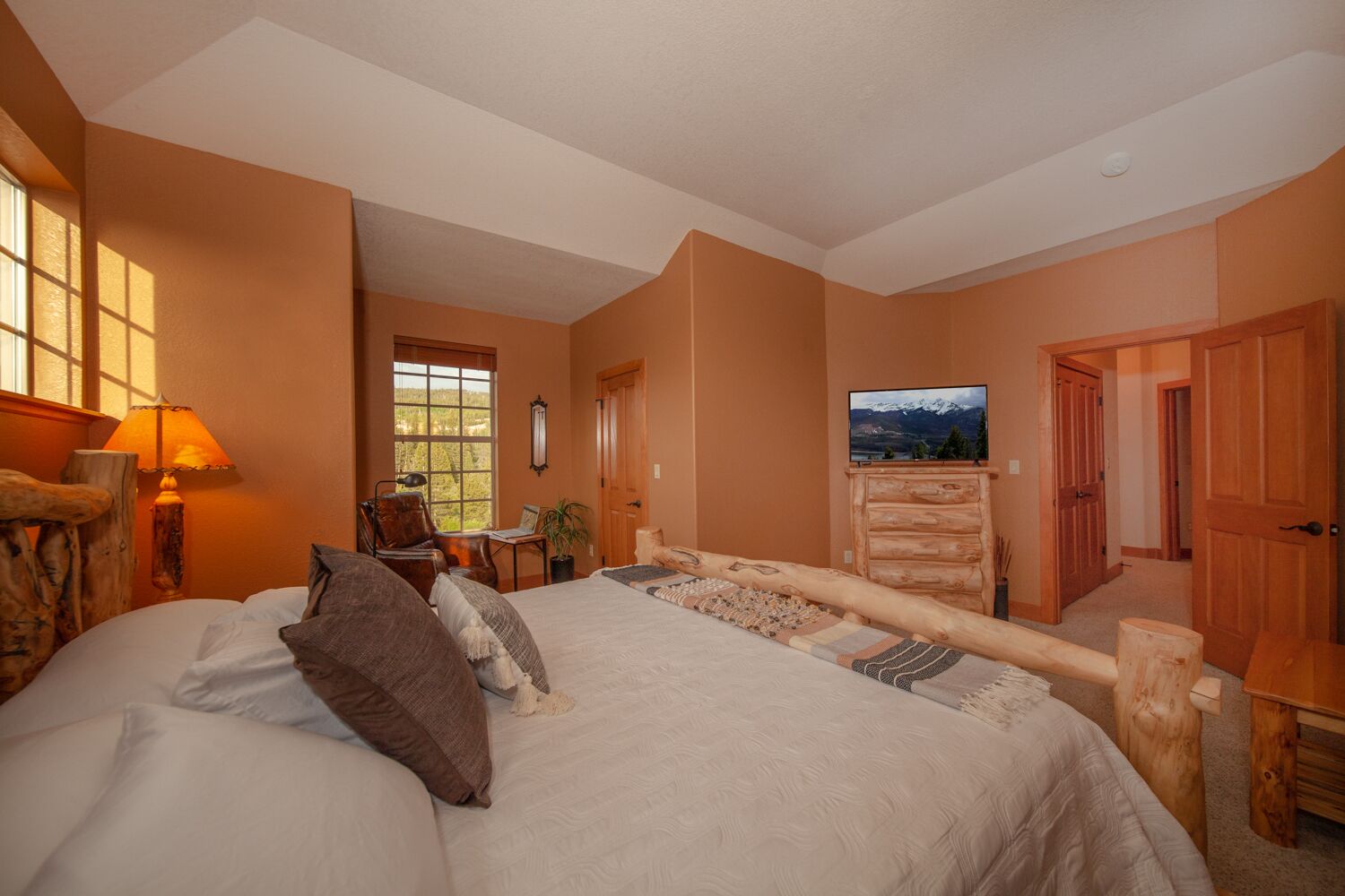 A bedroom with a large bed, a wooden dresser, a desk, and a chair. There is a window with a view of mountains.