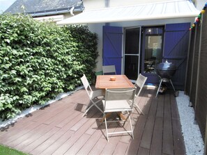 A patio with a wooden table and chairs, a barbecue grill, and a hedge.