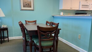 Un coin repas avec une table et des chaises en bois, un lustre, un tableau encadré au mur, et un espace cuisine avec des placards et un four à micro-ondes.