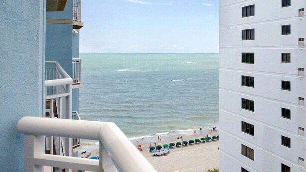 Un immeuble de grande hauteur doté de balcons donnant sur une plage où l’on aperçoit des parasols et des gens.