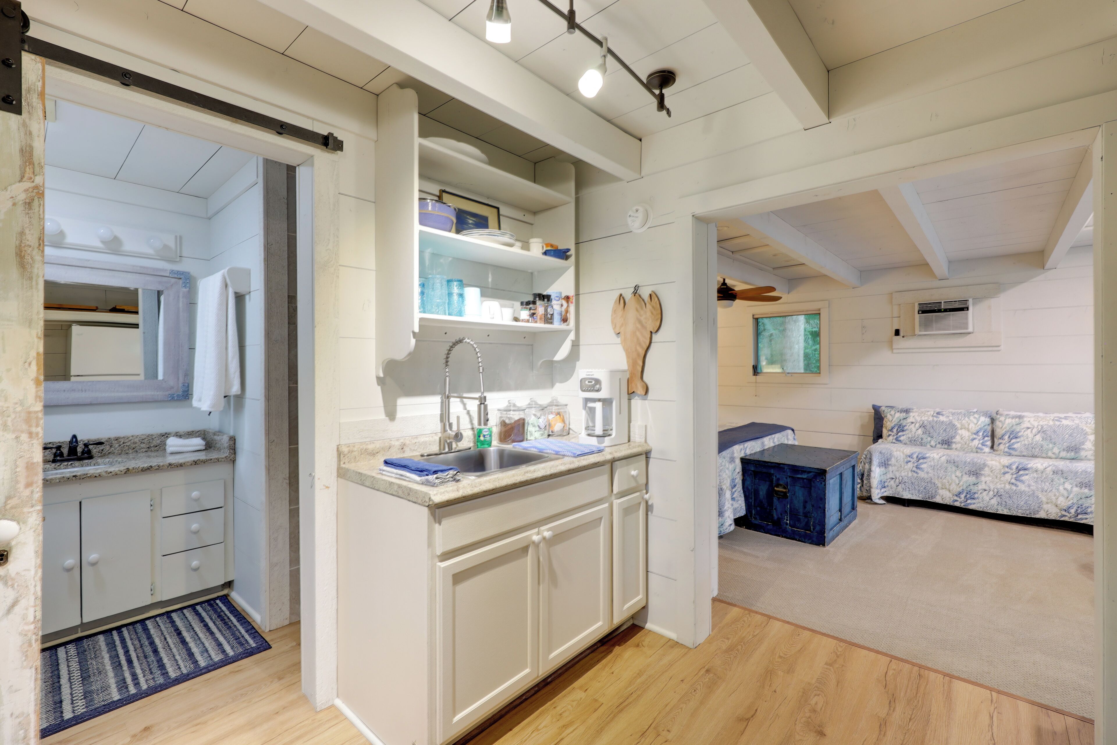 A modern kitchen with white cabinets, a built-in oven, and a large island with a sink. There is a wooden bench and a patterned sofa in the adjacent living area.