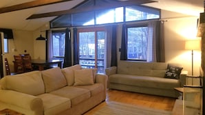 A cozy living room with a beige sofa, a grey armchair, and a wooden floor. Large windows with curtains provide natural light and a view of the outdoors. - Pocono Cabin in Masthope Mountain Community (Lackawaxen)