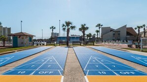 A shuffleboard court with blue and yellow markings, surrounded by palm trees and buildings.
