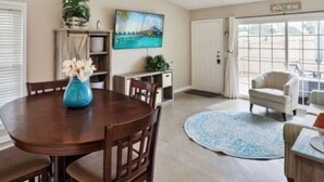A dining area with a round table, chairs, a ceiling fan, a TV, and a view through a glass door.