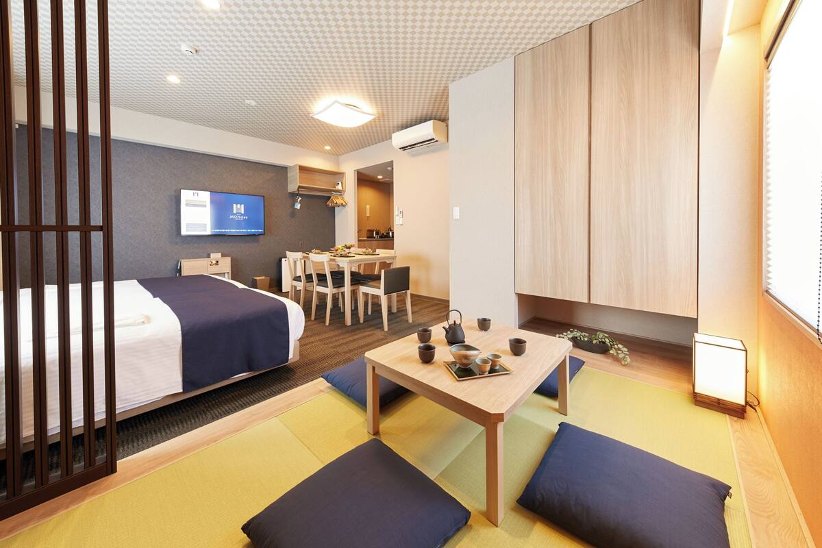 A hotel room with a bed, a desk with tea set, a TV, and a view of the kitchen.