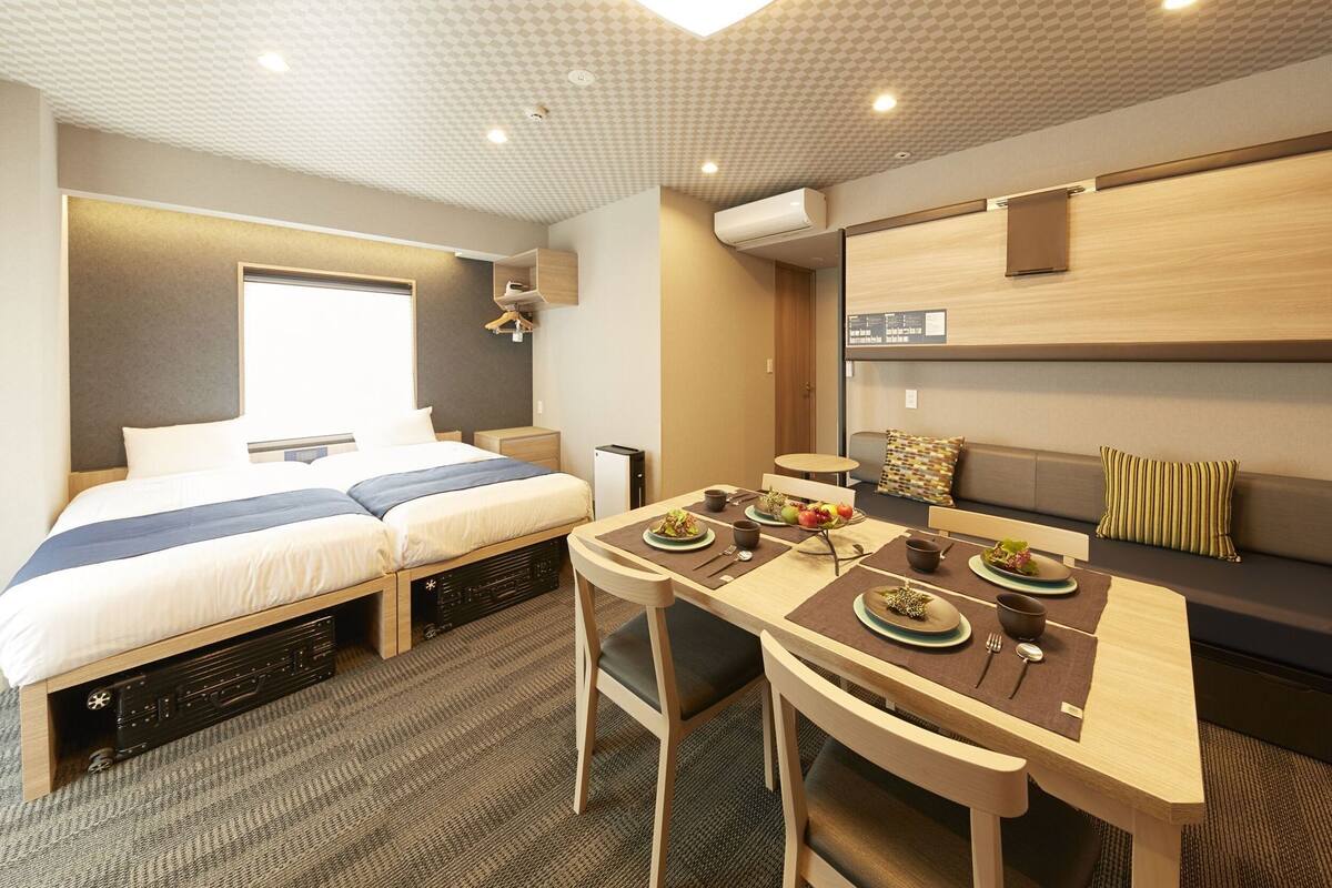 A hotel room with a bed, a dining table with chairs, a sofa, and an air conditioning unit.