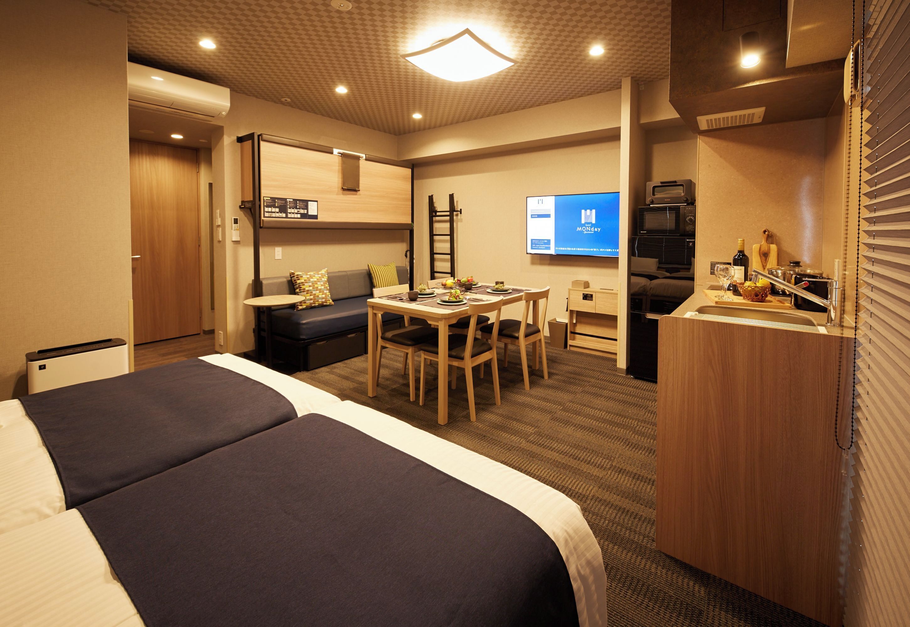A hotel room with a bed, a desk, a chair, a small table, a sofa, a TV, and a minibar.