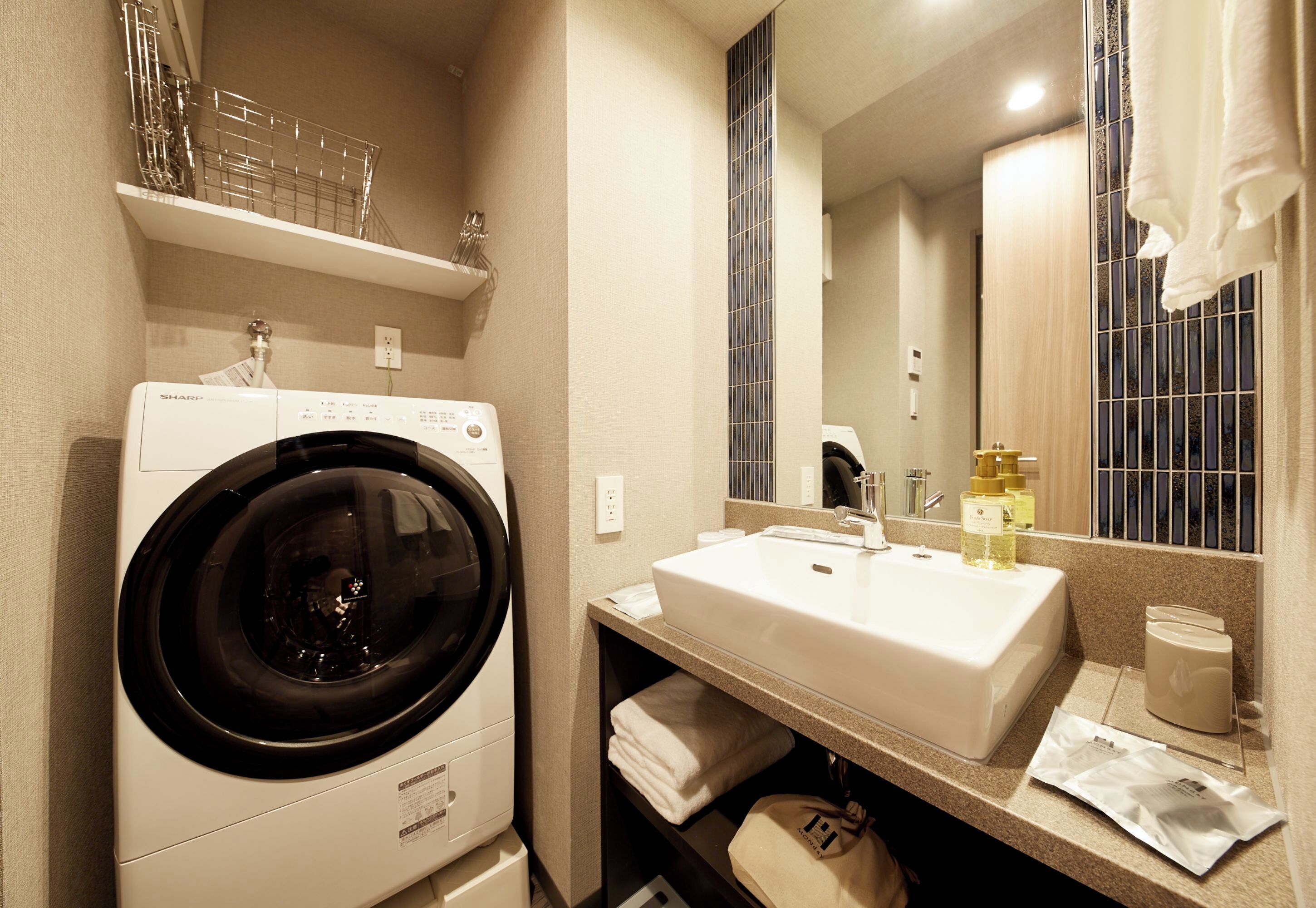 A compact hotel bathroom with a front-loading washing machine, a sink with a mirror above, and a towel rack.